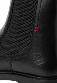 Botín de piel negra con un lateral elástico texturado en negro, costuras rojas y un logotipo grabado cerca de la suela. Material liso y duradero.