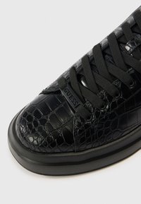 Mustad tenniskingad, millel on tekstuuriga krokodillinaha muster, ümar nina, mustad paelad ja kummist tald väikese aksendiga.