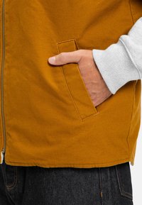 Gilet con zip realizzato in tessuto giallo senape, dotato di tasca laterale, con un polsino a costine grigio chiaro visibile al polso.