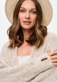 Beżowy, teksturowany kardigan z luźnym krojem, z szerokimi rękawami i małą brązową etykietą na mankiecie. Noszony na jasnym topie.