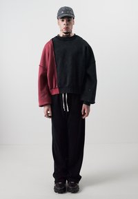 Übergroßer schwarzer und roter Pullover mit herunterhängenden Schultern, kombiniert mit schwarzen Hosen. Die Schuhe haben ein klobiges Design mit orangefarbenen Akzenten.