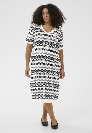 Žena s kučeravými vlasmi, oblečená v bielych šatách po kolená s krátkymi rukávmi, na ktorých sú čierne vlnité pruhy, a čiernych plochých topánkach, stojaca na bielom pozadí.