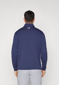 Μπλε σκούρο μπλουζάκι πόλο με μακριά μανίκια, γιακά και λευκό λογότυπο στο πάνω μέρος της πλάτης. Λιτή ύφανση και εφαρμοστή σχεδίαση. Δείχνονται ανοιχτό γκρι παντελόνια από κάτω.