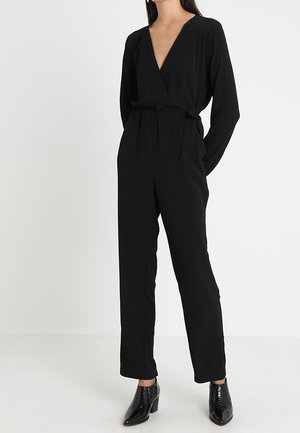 Sort jumpsuit med dyb V-hals, lange ærmer og bælte i taljen. Har lige ben og teksturerede, blanke sorte sko.