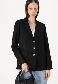 Blazer noir texturé avec six boutons en argent, deux poches avant et un revers cranté. Porté avec un jean bleu clair et un sac à main noir.