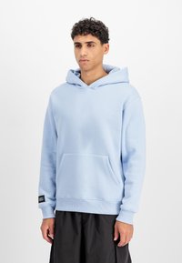 Világos kék kapucnis pulóver puha anyagból, elülső zsebből, húzózsinórral ellátott kapucnival, bordázott mandzsettákkal és fekete címkével a szegélyen.