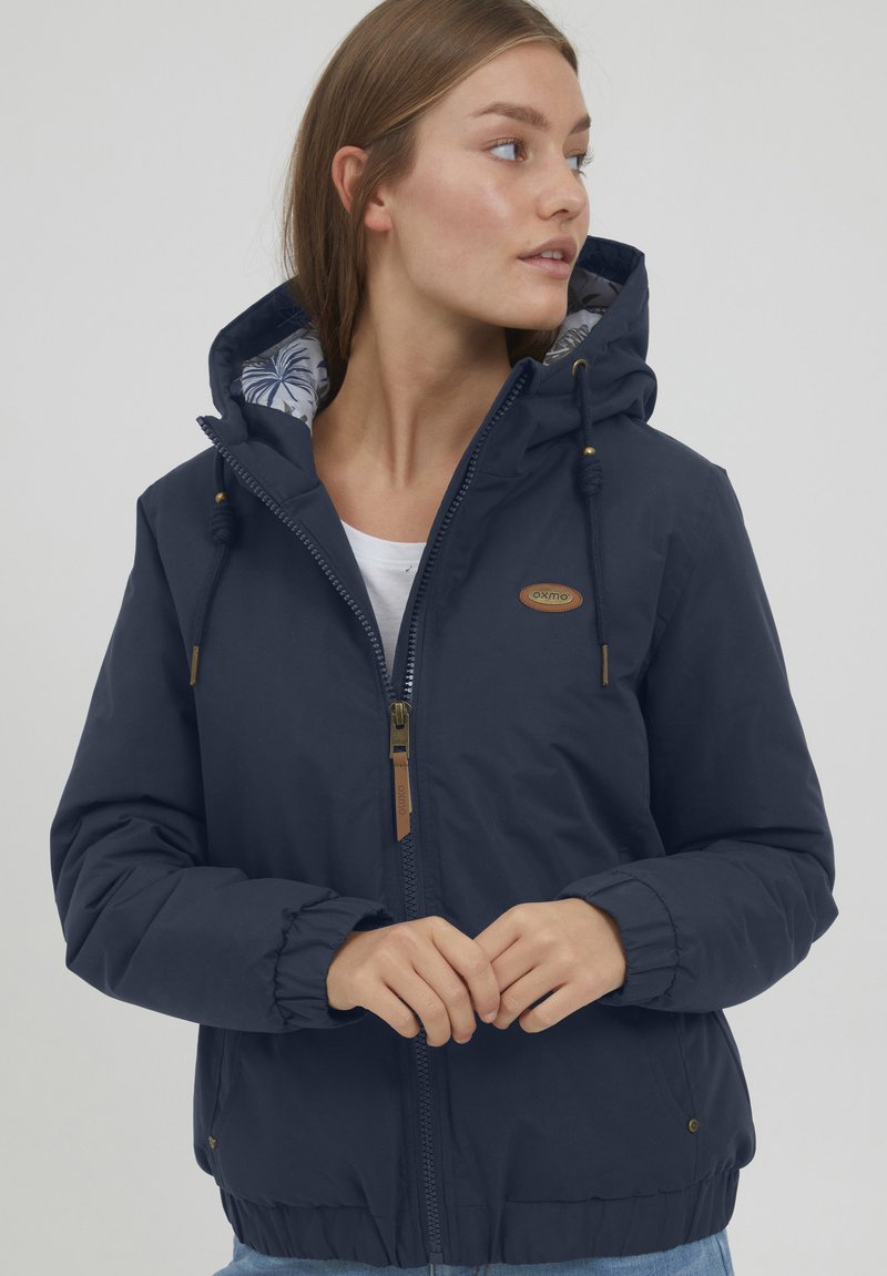Giacca con cappuccio blu navy, chiusura con zip, polsini elasticizzati e cordini. Foderata con un motivo floreale, presenta una patch con il logo sul petto.