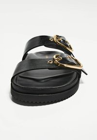 Svart sandal med två böjda remmar, prydd med guldbuckle-accenter och en texturerad sula. Slät läderfinish med en vadderad fotbädd.