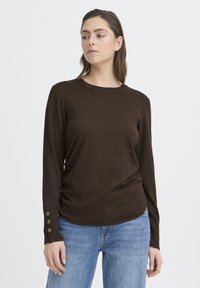 Pruun pikkade varrukatega top ümmarguse kaelusega, tekstuurse kangaga ja nööpdetailidega varrukaotsas; kombineeritud heleroheliste teksadega.