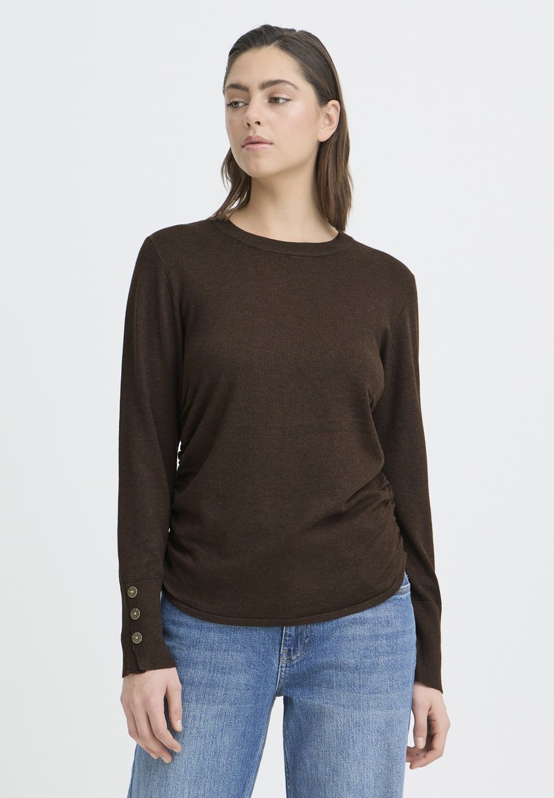 Pruun pikkade varrukatega top ümmarguse kaelusega, tekstuurse kangaga ja nööpdetailidega varrukaotsas; kombineeritud heleroheliste teksadega.