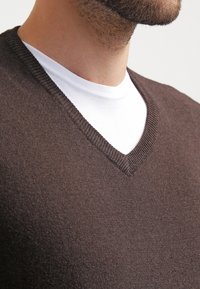 Brun ull v-ringad tröja lager på lager över en vit polotröja. Har ribbad halsringning, mjuk känsla och en kroppsnära passform runt halsen.