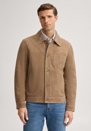 Uomo che indossa una giacca scamosciata marrone chiaro sopra una camicia a righe e jeans blu su uno sfondo chiaro uniforme.