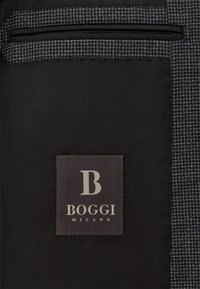 Črna tkanina z teksturiranim houndstooth vzorcem. Glavna oznaka prikazuje veliko črko "B" in "BOGGI MILANO" v sivi barvi na temnem ozadju.