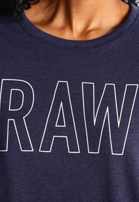 Laivasholta oleva puuvillapaita, jossa on suuri valkoinen teksti "RAW" rinnan kohdalla, sileä pinta ja klassinen pyöreä kaula-aukko.