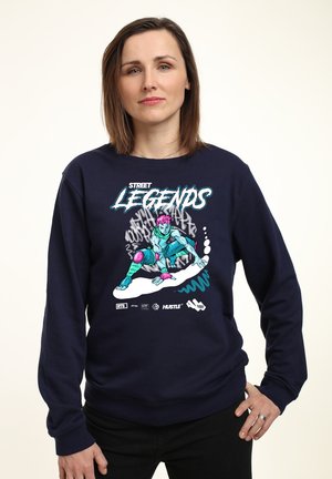 Sweatshirt bleu marine avec un motif graphique coloré représentant un skateur aux cheveux roses. Comprend le texte "LÉGENDES DE LA RUE" et des détails de logo.
