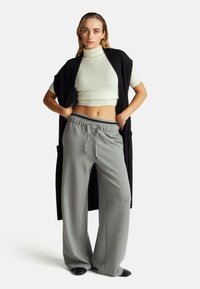 Frau in einem weißen, cropped Rollkragenpullover, schwarzem ärmellosem Cardigan und lockeren grauen Sweatpants mit Händen in den Taschen vor einem schlichten Hintergrund.