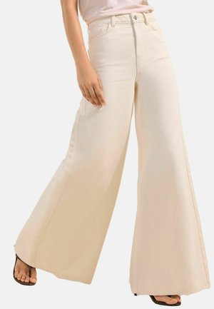 Žena nosiace krémové široké nohavice s vysokým pásom a viditeľnými prednými vreckami, čierne sandále na podpätku, kráča pred jednoduchým pozadím.