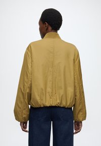 Żółta kurtka bomberka musztardowego koloru z gładką fakturą, elastycznym obszyciem dolnym i szerokimi rękawami. Prosty kołnierz i przeszycia z tyłu.