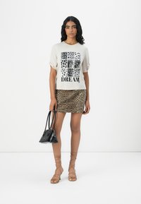 T-shirt beige à manches courtes avec un graphisme léopard et le texte "DREAM", associé à une jupe mini imprégnée de léopard, un sac à main noir et des sandales nues.