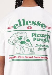 Bílý bavlněný tričko s zeleno-červeným grafickým designem. Tisk zobrazuje kus pizzy s houbami a textem "Pizzeria Perugia."