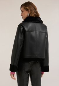 Donna con capelli castano chiaro che indossa una giacca nera in ecopelle con collo e polsini di pelliccia, pantaloni neri e orecchini argentati, rivolta verso sinistra.