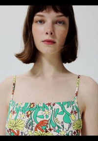 Jeune femme aux cheveux bruns courts portant un haut spaghetti coloré à motifs floraux, regardant directement la caméra sur un fond uni.