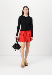 Čierny kardigán s dlhým rukávom, červená plisovaná mini sukňa s šnúrkou, béžové platformové clogy a čierna kvetinová taška s šnúrkou.