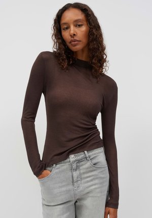 Vrouw met krullend haar, gekleed in een aansluitend donkerbruin lang mouwen shirt en lichtgrijze spijkerbroek, met één hand in de zak tegen een effen achtergrond.