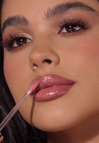 Un gloss à lèvres rose brillant appliqué sur des lèvres pleines à l'aide d'un applicateur en wand, avec un maquillage naturel et des sourcils définis. La peau a une finition douce et radieuse.