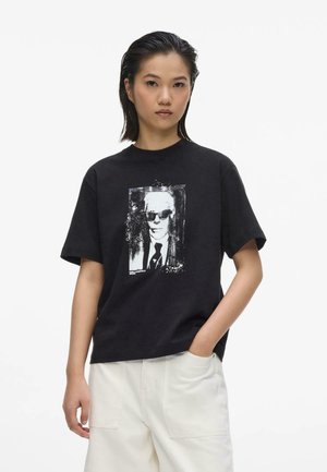 Ung kvinde med kort sort hår har en sort T-shirt med et sort-hvidt billede af en mand i solbriller og hvide bukser.