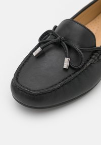 Mocassin en cuir noir avec un bout rond, détail de nœud sur le dessus, et accents en métal argenté avec des coutures visibles le long du bord.