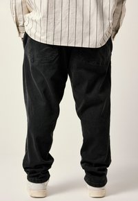 Pantalones negros holgados de denim, con bolsillos traseros y una superficie ligeramente texturizada. Combinados con zapatillas blancas en los tobillos.