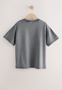 Camiseta gris de algodón de manga corta colgada en una percha de madera contra una pared blanco roto, vista desde atrás con detalles de costura visibles.