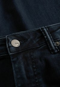 Donkere denim jeans met een metalen knoop waarop de merknaam "Gabba 1989" staat. Close-up van de tailleband en het stiksel.
