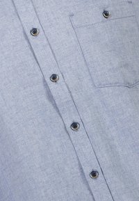 Camicia di chambray azzurro chiaro con fronte a bottoni, quattro bottoni scuri con accenti dorati e taschino sul lato sinistro.