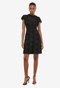 Rochie neagră din dantelă cu mâneci scurte bufante, model floral, guler înalt și siluetă conică. Asortată cu sandale negre cu bretele.