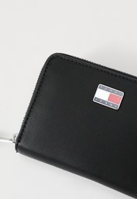 Crni kožni novčanik na zip s teksturiranom površinom. Ima srebrnu logotip oznaku s plavim, bijelim i crvenim naglascima. Minimalistički dizajn.