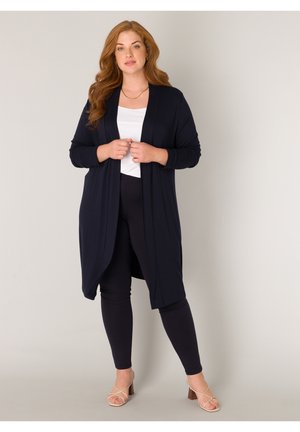 Vrouw met lang rood haar, een wit shirt, een zwarte lange cardigan, zwarte broek en beige sandalen met hak, staand tegen een effen achtergrond.