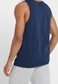 Laivastonsininen toppi, jossa on pyöreä pääntie ja pieni logotunnus takana. Kangas näyttää pehmeältä ja kevyeltä.