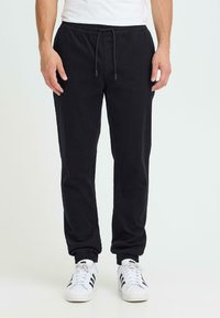 Pantalon de survêtement noir avec une taille élastique, un cordon de serrage et des poignets ajustés. Texture de tissu douce, associé à des chaussures de sport blanches.