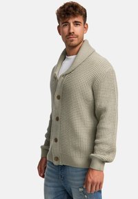 Cardigan de punto gris claro con cuello chal, botones de madera y puños acanalados, llevado sobre una camiseta blanca y combinado con jeans azul claro.