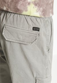 Pantaloni cargo gri deschis dintr-un material durabil, cu un buzunar pe spate cu clapetă și o etichetă de marcă mică. Talie elasică pentru comfort.