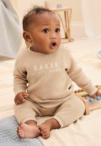 Ensemble en tricot beige avec des poignets côtelés, arborant le texte de logo blanc "BAKER BY TED BAKER". Le bébé est assis sur une couverture bleue texturée, tenant un jouet en bois.