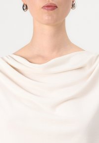 Cremig drapierter Stoffoberteil, weiche Textur, Cowl-Ausschnitt, mit enganliegendem Design am Hals, sichtbar mit silbernen Creolen.