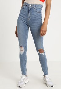 Nainen, joka on pukeutunut vaaleansinsisiin korkeavyötäröisiin skinny-farkkuihin, joissa on revittyjä polvia, ja valkoisiin lenkkareihin, seisoo yksinkertaista taustaa vasten.