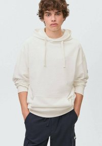 Krämliknande hoodie i mjukt tyg, med en känguruficka, snörning och ribbade muddar. Bärs med mörka byxor.