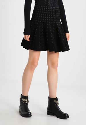 Femme portant une jupe évasée noire à motif à pois et des bottines noires avec des boucles dorées avec le logo "Versace Jeans".