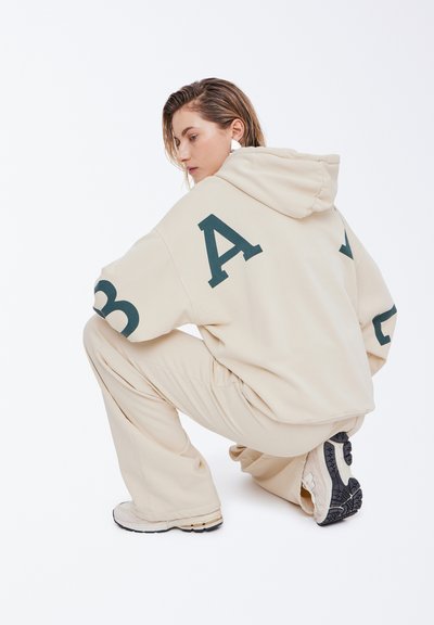 Beige hettegenser med store grønne bokstaver, paret med matchende beige bukser og lyse sneakers, med en teksturert overflate.