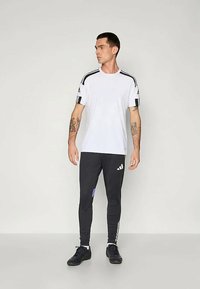 Balta īsām piedurknēm krekls ar melnām svītrām uz pleciem, apvienots ar melnām legingām ar violetu akcentu un baltu logotipu, valkāts ar melnām kurpēm.