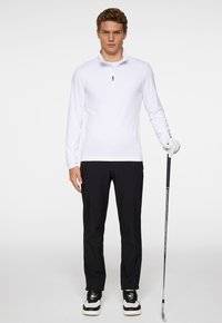 Vit långärmad tröja med halvspringa och krage, ihop med svarta byxor och svartvita golfskor. Håller en golfklubba.